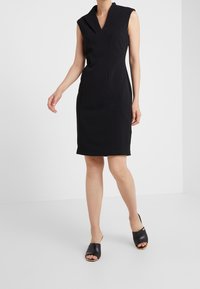 Robe noire sans manches avec un décolleté en V profond, une silhouette ajustée et des détails de couture visibles. Associée à des mules noires à bout ouvert.