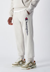 Pantalon de survêtement gris clair Champion avec un logo bleu marine et le texte "Champion". Caractéristiques : coupe décontractée et tissu doux, associé à des baskets multicolores.