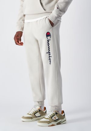 Pantalones de chándal de color gris claro de Champion con un logotipo en azul marino y el texto "Champion". Presentan un ajuste holgado y una tela suave, combinados con zapatillas multicolores.