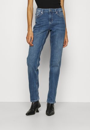 Vrouw draagt mid-rise blauwe recht gesneden jeans met voorzakken en zwarte enkellaarsjes, staand tegen een effen lichte achtergrond.