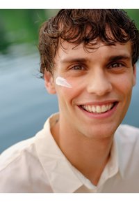 Crème appliquée sur la joue d'un jeune homme souriant avec des cheveux courts et bouclés de couleur brune, portant une chemise claire, avec de l'eau en arrière-plan.