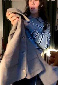Vrouw houdt een beige jas vast, draagt een gestreept shirt met lange mouwen, staat binnen met zonlicht dat door een raam naar binnen valt.