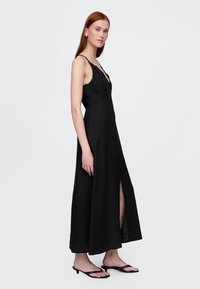 Schwarzes Maxikleid mit dünnen Trägern, Spitzenbesatz am Ausschnitt, einem vorderen Schlitz und einem taillierten Oberteil. Kombiniert mit schwarzen flachen Sandalen.