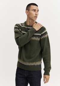Grüner Strickpullover mit einem hohen Kragen, der ein geometrisches Muster in Creme, Schwarz und Braun über die Brust und den Saum aufweist. Gerippte Bündchen.