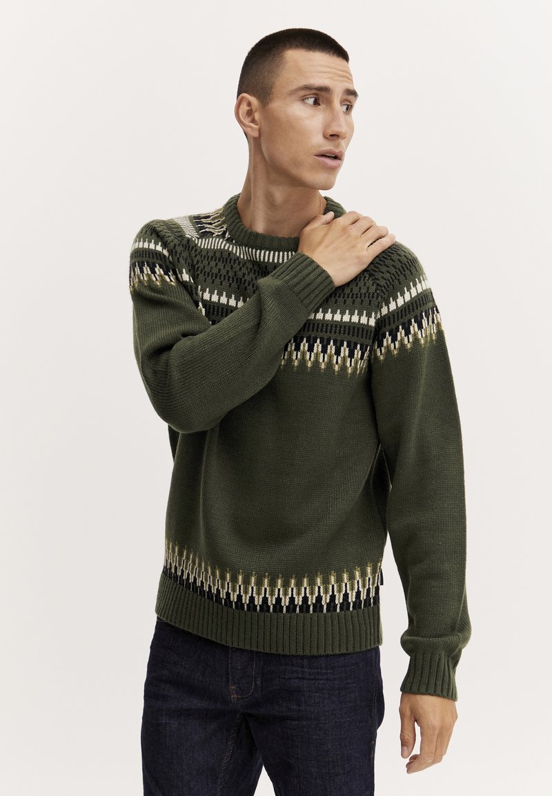 Grüner Strickpullover mit einem hohen Kragen, der ein geometrisches Muster in Creme, Schwarz und Braun über die Brust und den Saum aufweist. Gerippte Bündchen.