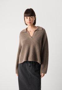 Pull en maille texturée marron avec un décolleté en V et un col, doté de manches larges. Assorti avec une jupe en denim noire. Coupe légèrement cropped.