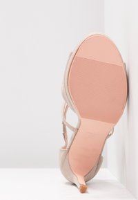 Chaussure à talon haut en daim beige avec un bout pointu et une semelle rose, présentée debout contre un mur d'angle blanc.