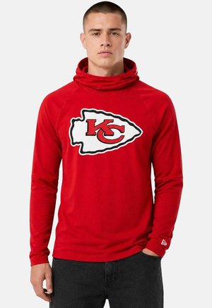 Uomo che indossa una felpa rossa con cappuccio e logo dei Kansas City Chiefs sul petto e pantaloni neri, in piedi con una mano in tasca.