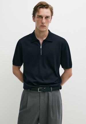 Giovane uomo con capelli castano chiaro che indossa una polo nera a maniche corte con mezza cerniera e pantaloni grigi plissettati con cintura nera.