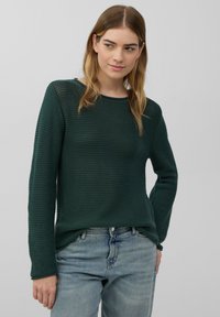 Grüner strukturierter Pullover mit langen Ärmeln und Rundhalsausschnitt, kombiniert mit hellblauen Jeans. Das Material wirkt gewebt mit einem groben Muster.