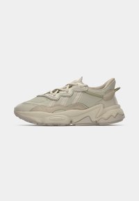 Sneaker beige chiaro con materiali in mesh testurizzato e suede, con una suola spessa, linee curve e lacci sulla parte superiore per supporto.