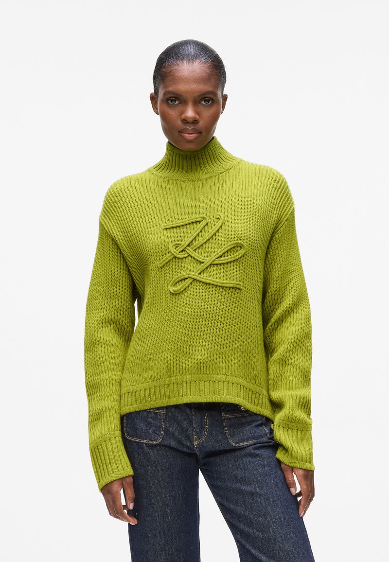 Grüner gerippter Strickpullover mit hohem Kragen, der ein erhabenes Logo-Design auf der Vorderseite und übergroße Ärmel aufweist, kombiniert mit dunklen Jeans.