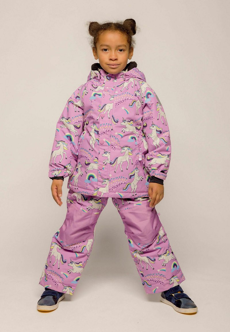 Enfant en combinaison de pluie violette avec des motifs de licorne et d'arc-en-ciel, debout jambes écartées et mains détendues le long du corps.