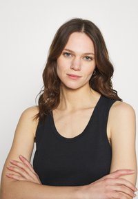 Top senza maniche nero con scollatura rotonda, realizzato in tessuto morbido. Esposto su un modello con sfondo neutro, braccia incrociate.