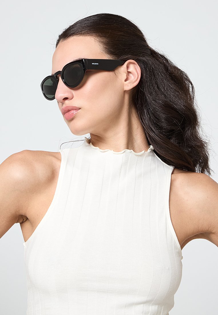 Des lunettes de soleil noires avec une monture géométrique et des verres foncés, portées par une personne ayant de longs cheveux noirs, sur un fond uni.