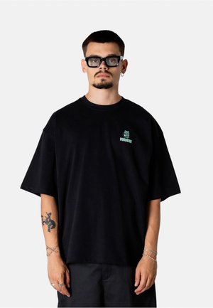 Mand med kort hår og skægstubbe iført sort oversized Woodbird-t-shirt, sorte briller, sølv hoop-ørering, tatoveringer og armbånd mod en ensfarvet baggrund.
