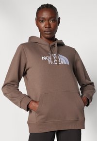 Hoodie castanho com um bolso frontal, apresentando um grande logótipo branco. Feito de um tecido suave com um corte descontraído e capuz ajustável com cordão.