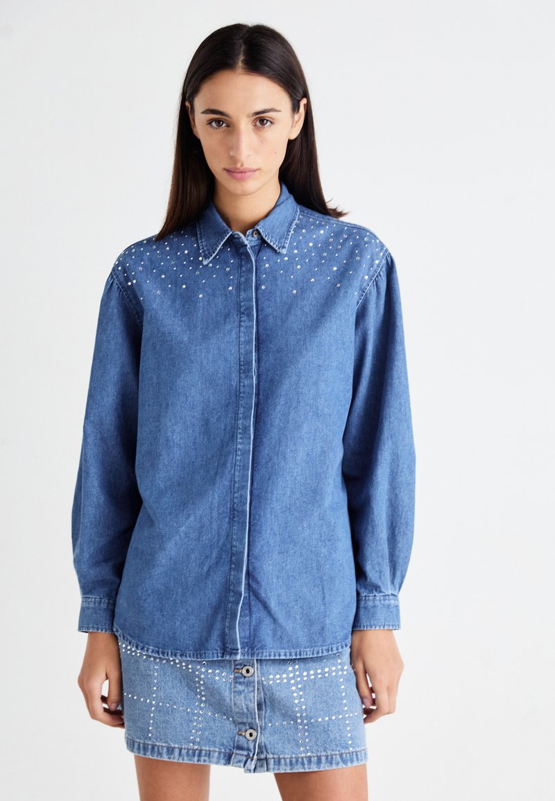 Denim shirt with a button-down collar, long sleeves, and studded embellishments on the shoulders. Paired with a matching denim skirt.