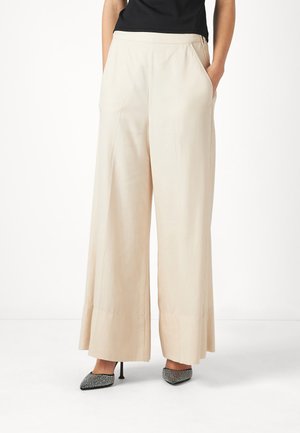 Pantaloni a gamba larga in tessuto beige leggero, con tasche laterali e una cintura liscia, abbinati a tacchi a punta neri e bianchi con motivo.