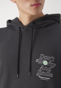 Felpa grigia scura con cappuccio e cordini, con una grafica frontale che riporta il testo "Non Preoccuparti e Sorridi" accanto a una faccina sorridente verde.
