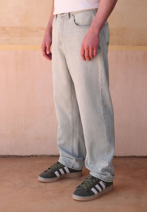 Ein Mann trägt hellblaue Jeans, ein weißes Hemd und dunkelgrüne Adidas-Sneaker und steht auf einem beigen Boden vor einer beigen Wand.