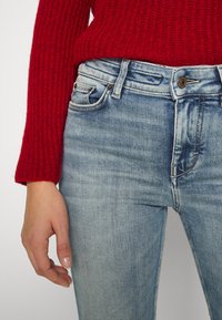 Vaqueros de mezclilla azul claro con un diseño desvanecido, estilo de cinco bolsillos y cierre de botón metálico, combinados con un suéter de punto acanalado rojo texturizado.