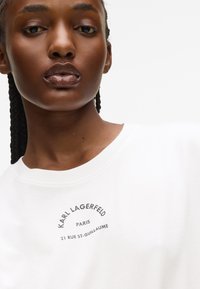 Zbliżenie na osobę z warkoczami, ubraną w białą koszulę z napisem "KARL LAGERFELD PARIS 21 RUE ST-GUILLAUME" na piersi.