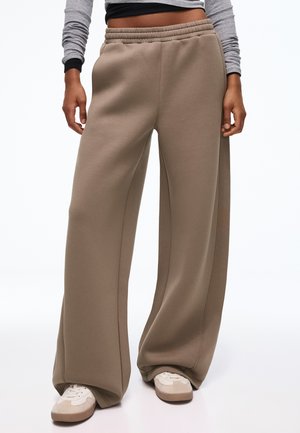 Personne portant un pantalon beige à jambes larges, des baskets blanches et un haut gris à manches longues avec des poignets noirs, debout devant un fond uni.