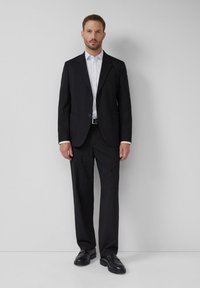 Homme portant un costume noir, une chemise blanche avec de petits pois noirs, une ceinture noire et des chaussures en cuir noires, se tenant contre un mur gris clair uni.