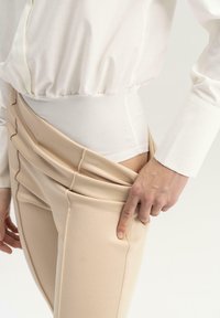 Los pantalones ajustados beige presentan una textura suave, un diseño plisado y bolsillos laterales. Se lleva una blusa blanca, con mangas recogidas y puños.