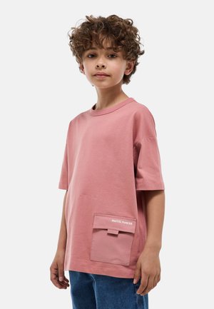 Enfant aux cheveux bouclés portant un t-shirt rose surdimensionné avec une poche "PASTEL POWER" et un jean bleu, debout devant un fond uni.