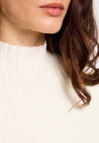Parte inferiore del viso di una donna con labbra lucide, capelli castani mossi, che indossa un dolcevita bianco a coste.