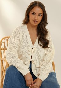 Krämfärgad stickad cardigan med ett diamantmönster, med en djup V-halsringning och två framknytningar. Oversized ärmar och en avslappnad passform.