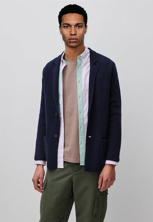 Joven con americana azul marino texturizada sobre camiseta beige en capas y camisa verde claro abotonada, con pantalones cargo color oliva.