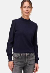 Pull bleu marine avec un col montant et des manches longues côtelées, présentant un motif texturé et une silhouette ajustée. Associé à un jean gris.