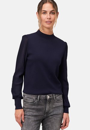 Pull bleu marine avec un col montant et des manches longues côtelées, présentant un motif texturé et une silhouette ajustée. Associé à un jean gris.