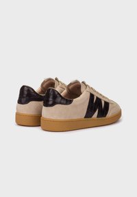 Zapatillas de ante beige con acentos texturizados en negro, con cordones planos, suela de goma y un logo en el costado. La vista trasera muestra materiales contrastantes.