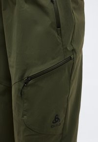 Õllerohelised välispüksid kahe nähtava lukuga taskuga ja must Odlo logo põlve lähedal.