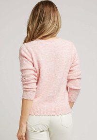 Pull en doux rose clair, duveteux, avec des poignets et un ourlet côtelés, présentant des détails en forme de vagues. Tissu texturé et coupe ajustée.