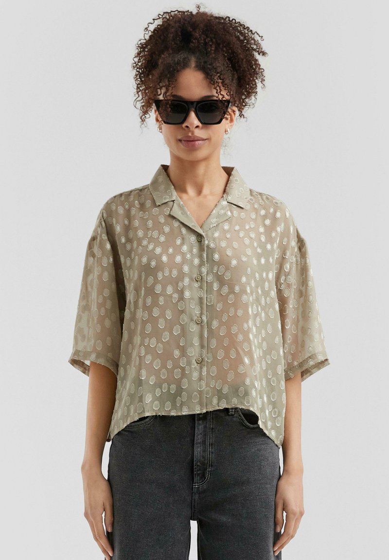 Camicia verde chiaro a maniche corte con un tessuto trasparente e un motivo a pois in rilievo. Presenta un colletto classico e chiusura con bottoni frontali.