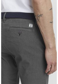 Grå vävda byxor med en texturerad yta, featuring en bakficka med en knapp och en liten varumärkesetikett. En elastisk marinblått bälte är synligt.