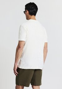 Camicetta bianca a maniche corte con motivo testurizzato a forma di foglia di palma, abbinata a pantaloni corti verde oliva, vista da dietro.