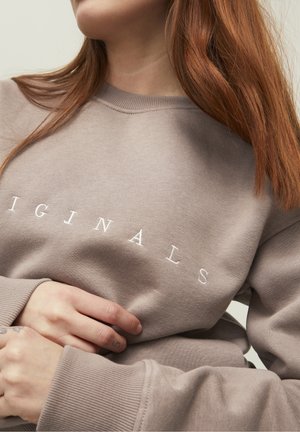 Jeune personne aux longs cheveux roux portant un sweat-shirt taupe avec « ORIGINALS » imprimé en blanc sur la poitrine, mains jointes à la taille.