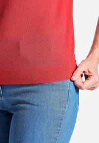 Rode korte mouwen trui met een geribde zoom, gecombineerd met blauwe denim jeans. Detail van een hand die de zijkant van het shirt bij de zoom vasthoudt.