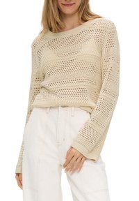Pull beige à maille ouverte avec une coupe décontractée, présentant des motifs horizontaux texturés et des manches larges, associé à un pantalon de couleur claire.
