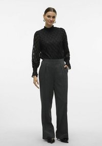 Blusa nera trasparente con motivo testurizzato e colletto alto, abbinata a pantaloni grigi a gamba larga su misura. Il modello indossa grandi orecchini d'argento.