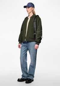 Jacket bomber verde oliva com fecho, camisola de malha bege, jeans azul claro e sapatos de plataforma pretos. Boné azul com padrão de corações.