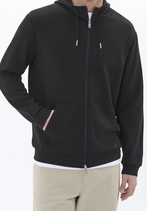 Man wearing a black zip-up hoodie with front pockets, beige pants, and a white undershirt against a plain background.