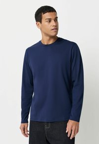 Cămașă navy cu mânecă lungă dintr-un material neted, cu gât rotund. Prezintă o cusătură minimă și un tiv drept, asortată cu pantaloni din denim închis.