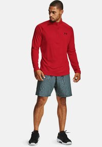 Rød langærmet atletisk top med halv-zip design, parret med grå mønstrede shorts. Sorte sneakers fuldender outfittet.